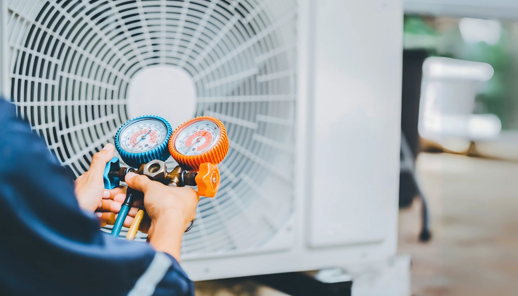 A technician holds blue and orange pressure gauges in front of an air conditioning unit, ready for maintenance or repair, ensuring optimal performance while managing AC recharge costs.