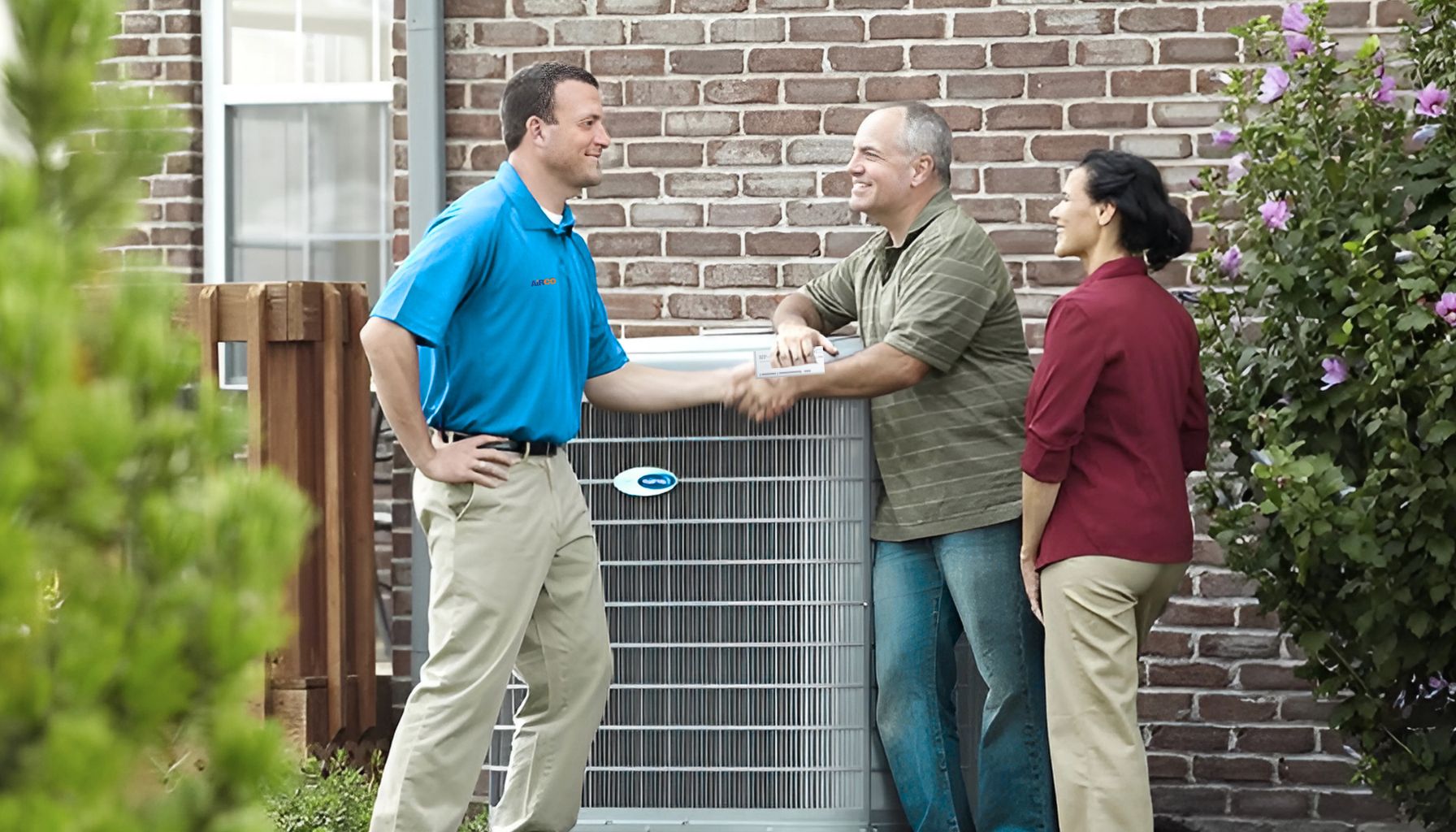 A technician in a blue shirt shakes hands with a homeowner outside a house, discussing an air conditioning unit and the potential tax rebate on AC repair units. A woman observes nearby.