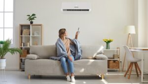 A woman wrapped in a gray blanket sits on a beige couch, converting an air conditioner ac to heat, surrounded by plants and modern decor.